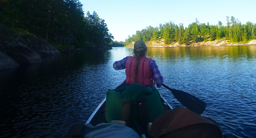From the back of a canoe, you can see the person in front paddling. The boat is on calm water with trees lining the shore. 